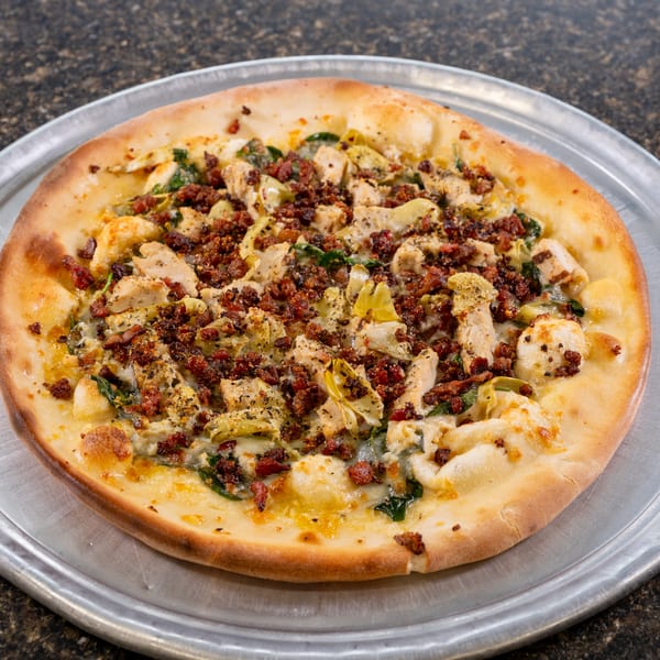 A whole pizza with chicken, bacon, artichoke hearts, and spinach on a metal tray.