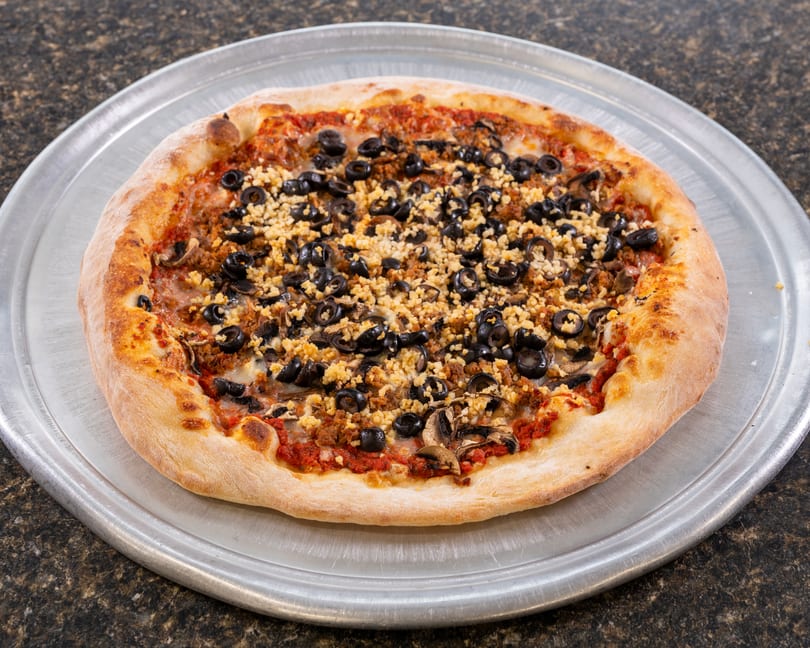 A whole pizza with black olives, crumbled sausage, and mushrooms on a metal pan.