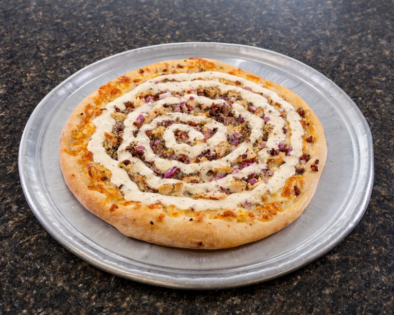 A round pizza with a spiral of white sauce, chicken, and red onions on a metal tray.