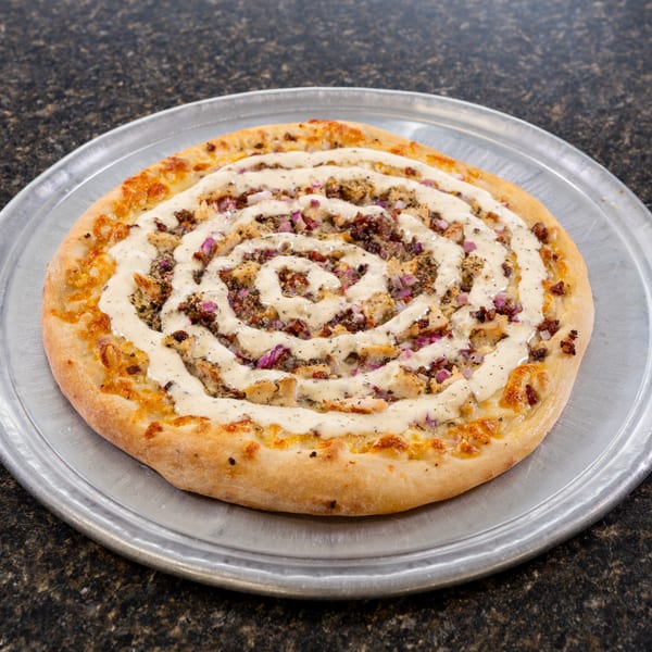 A round pizza with a spiral of white sauce, chicken, and red onions on a metal tray.