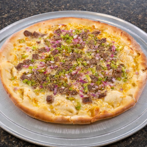 A whole pizza with white sauce, steak, green peppers, and red onions on a metal pan.