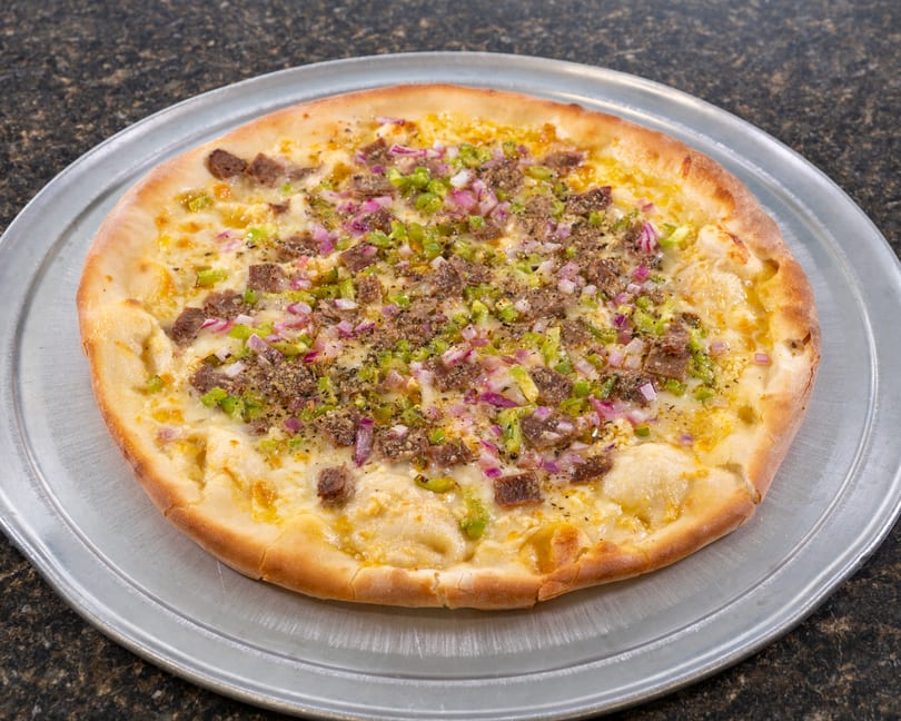 A whole pizza with white sauce, steak, green peppers, and red onions on a metal pan.