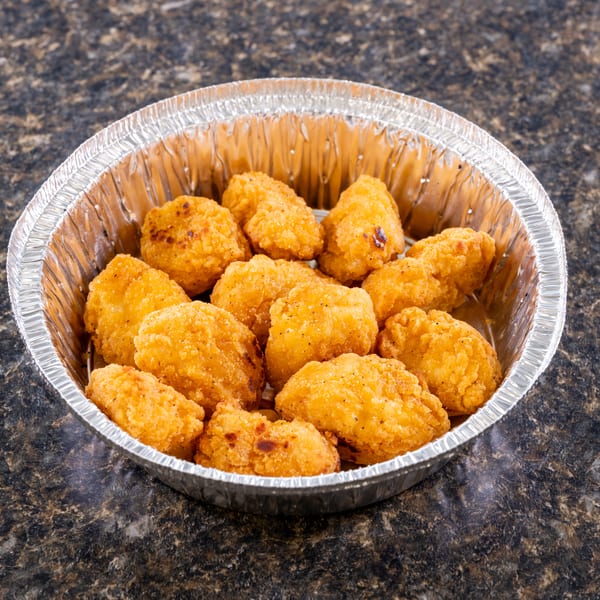 Golden-brown fried chicken nuggets in a round aluminum foil container on a dark speckled surface.