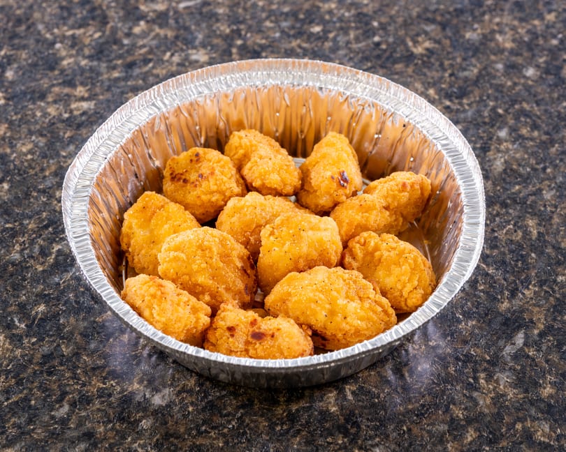 Golden-brown fried chicken nuggets in a round aluminum foil container on a dark speckled surface.
